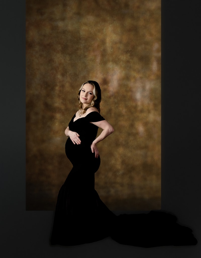 Photoshoot for maternity in studio. Black background with a gold canvas behind mom wearing a black dress.