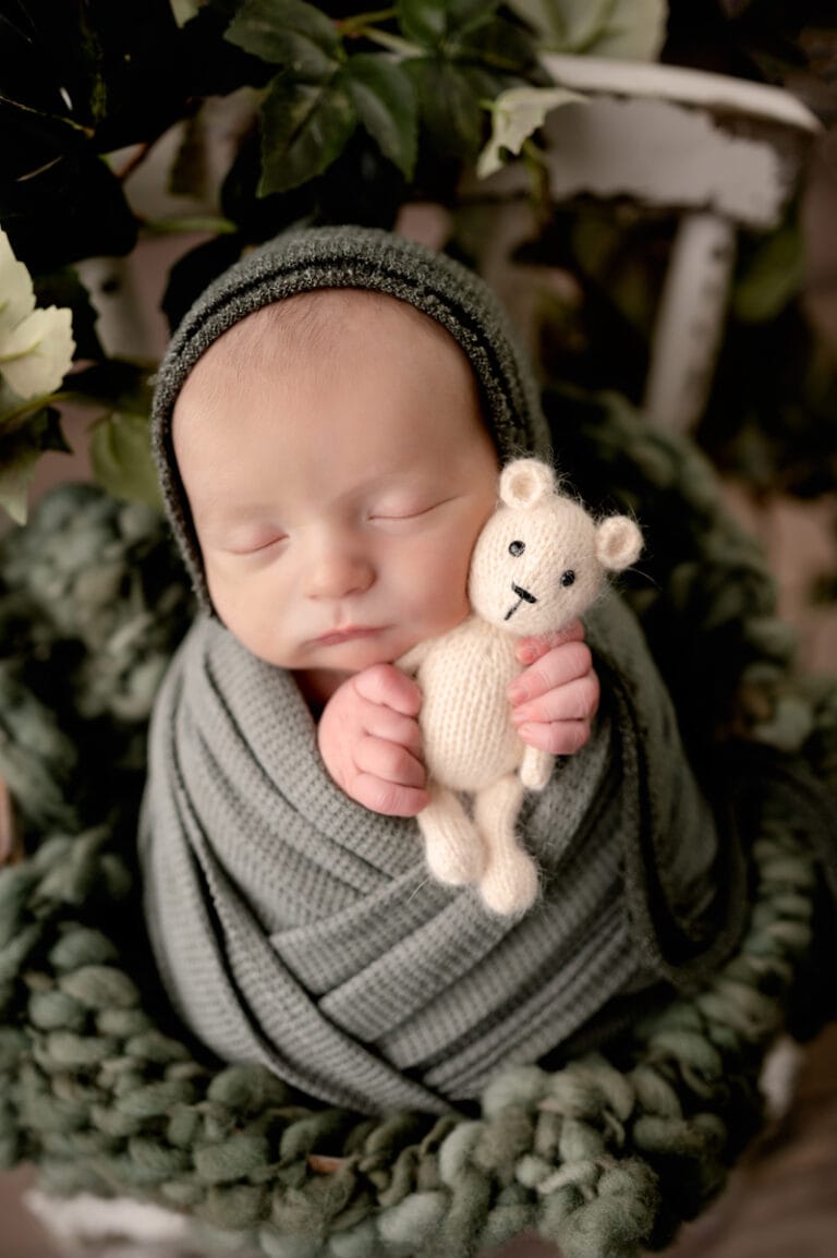 Newborn boy wearing a bonnet and holding a stuffed bear.