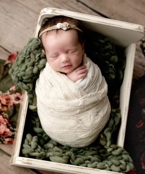 Newborn baby girl in a cream crate with a green fluffy under her. She is wrapped in a cream lace wrap and a flower layer is under the crate.