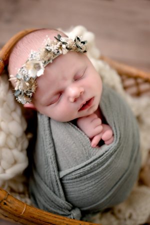 Newborn girl swaddled in a sage green wrap inside of a basket with a flower crown on her head.