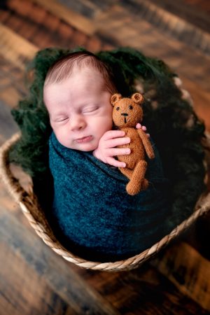 Newborn boy in a basket, holding a teddy bear, wrapped in a green wrap. Mor